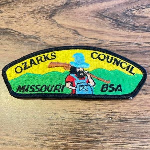 May include: An embroidered patch with a black border featuring a yellow sky and green hills. It depicts a figure in a blue hat, red shirt, and blue overalls carrying a brown rifle. Text reads OZARKS COUNCIL, MISSOURI, and BSA in black lettering.