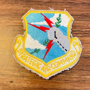 May include: Embroidered patch with a shield design. The patch features a light blue sky with white clouds, red lightning bolts, and a white hand holding a green sprig. The text "STRATEGIC AIR COMMAND" is in yellow.