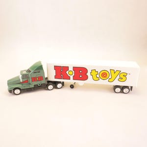 May include: A green toy semi-truck with a white trailer featuring the red and yellow K-B toys logo on the side. The truck cab has the letters K-B in red on the door. The vehicle has black wheels and a white trailer with a red and yellow logo.