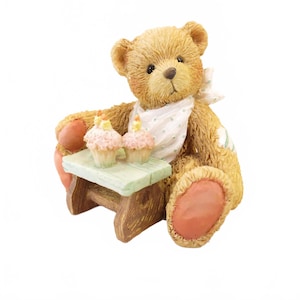May include: A brown teddy bear figurine with a white bib and a small green table. Two cupcakes with pink frosting and yellow candles sit on the table. The bear has a patch on its arm and pink pads on its feet.