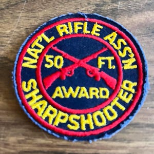 May include: A circular embroidered patch with a dark blue background and a red border. It features yellow text reading NATL RIFLE ASSN SHARPSHOOTER, with 50 FT. and two crossed red rifles in the center. The patch has a stitched edge finish.