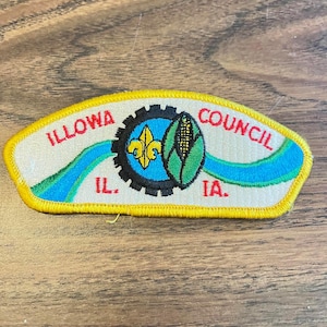 May include: Embroidered patch with the text "ILLOWA COUNCIL" in red, arched over a central design. The design includes a fleur-de-lis, a corn stalk, and a gear. The patch also features the abbreviations "IL." and "IA."