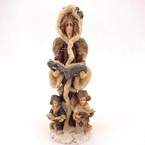 May include: A figurine of a woman and two children singing carols. The woman wears a burgundy coat with a fur-lined hood and holds a book. The children are in similar coats and also hold books. The base is textured to resemble snow.