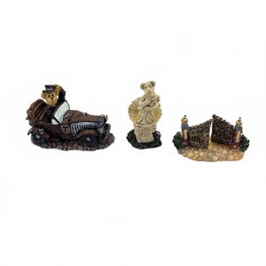 May include: Three miniature figurines: a vintage car with a bear driver, a bear statue in a shell, and a decorative gate. The car is brown and black, the statue is beige, and the gate is brown with gold accents. These are likely decorative items.