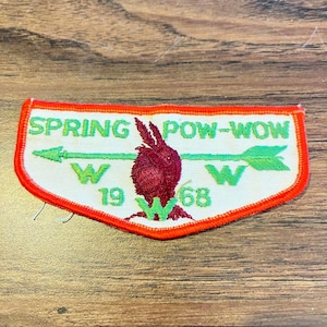 May include: A white embroidered patch with a bright orange border. It features the green text SPRING POW-WOW, a horizontal green arrow, the letter W on both sides, and the year 1968. A dark red hand gesture is centered in the middle of the patch.