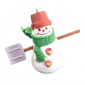 May include: A snowman ornament with a red bucket hat, green scarf, and two apple buttons. The snowman holds a wooden shovel and a wooden arm. The ornament is white with a smiling face and is ready for the winter holidays.