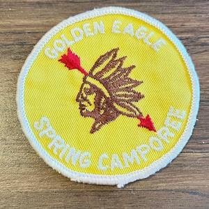 May include: Round yellow patch with white stitching and text. The patch features a Native American profile with a feathered headdress, pierced by a red arrow. The text reads "GOLDEN EAGLE SPRING CAMPOREE".