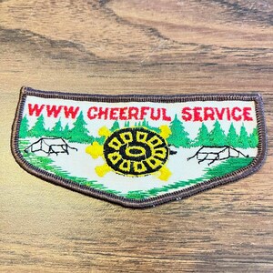 May include: A curved embroidered patch with a brown border featuring the red text WWW CHEERFUL SERVICE. The design includes green pine trees, two black-outlined tents, and a central yellow and black turtle graphic on a white background.