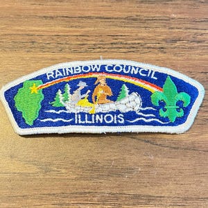May include: An embroidered patch with a blue background featuring a green map of Illinois, a rainbow, a canoe with two figures, and a green fleur-de-lis. The text reads RAINBOW COUNCIL and ILLINOIS in white capital letters with a white border.