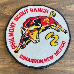 May include: Round patch with a red, yellow, and black bull illustration. The text "PHILMONT SCOUT RANCH CIMARRON, NEW MEXICO" is embroidered in red around the white background. A black border surrounds the patch.