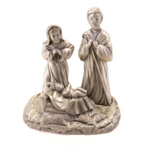May include: A silver-colored nativity scene depicting Mary, Joseph, and baby Jesus in a manger. The figures are detailed and stand on a textured base, creating a religious holiday decoration.