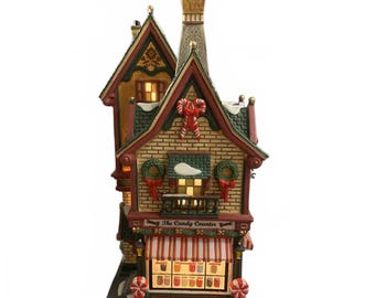 May include: A Christmas-themed ceramic candy shop with a red and white striped awning. The building has a brick facade, green roof, and is decorated with wreaths, bows, and snow. The sign reads "The Candy Counter".