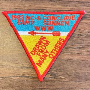 May include: A triangular patch with a red border, featuring text and an arrow design. The top section is blue with white text, the middle is white, and the bottom is yellow with red text. The text includes "1983 NC-5 CONCLAVE CAMP SUNNEN WWW" and "DRAWN FROM MANY OTHERS".