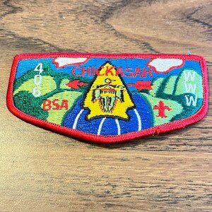 May include: Embroidered patch with a red border, featuring the text "CHICKASAH" in red, a yellow triangle, and the letters "BSA". The patch has a blue, green, and yellow color scheme, with white clouds and green hills.