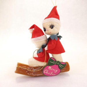 May include: A vintage Christmas ornament featuring two elf figures in red outfits and hats, riding on a wooden sled. The ornament has a pink tag with a green bow and the words "Merry Christmas".