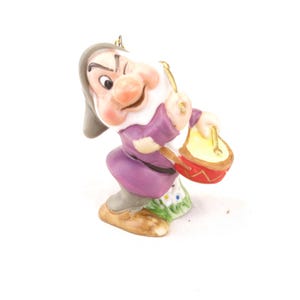 May include: A ceramic figurine of a character with a gray hat, purple tunic, and tan shoes, playing a small red drum with yellow drumsticks. The character has a winking expression, a large nose, and stands on a base with green grass.
