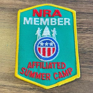May include: A shield-shaped embroidered patch with a green background and yellow border. It features the red text NRA MEMBER and AFFILIATED SUMMER CAMP. The center displays a white pine tree, two figures, and a circular emblem with stars and stripes.
