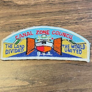 May include: An embroidered patch with a light blue sky and yellow land background. It features a ship in the center between orange lock gates. Text reads CANAL ZONE COUNCIL, THE LAND DIVIDED, THE WORLD UNITED, and B.S.A. in red, blue, and white thread.
