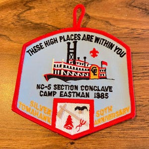 May include: A light blue and red embroidered patch with the text "THESE HIGH PLACES ARE WITHIN YOU" and "NC-5 SECTION CONCLAVE CAMP EASTMAN 1985". The patch features a steamboat and other graphics.