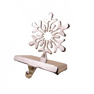 May include: Silver-colored metal stocking holder featuring a snowflake design. The snowflake is attached to a rectangular base with a hook for hanging. A festive Christmas decoration.