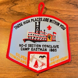 May include: A light blue embroidered patch with a red border and a hanging loop. It features a steamboat illustration, a red fleur-de-lis, and text reading "THESE HIGH PLACES ARE WITHIN YOU", "NC-5 SECTION CONCLAVE CAMP EASTMAN 1985", "SILVER TOMAHAWK", and "50TH ANNIVERSARY".