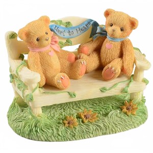 May include: A decorative figurine featuring two teddy bears seated on a cream-colored bench. The bears are light brown with pink accents and wear ribbons. The bench has a banner that reads "Heart to Heart". The base is green with flowers.