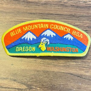 May include: An embroidered patch featuring a mountain range with white peaks against a blue sky and orange background. The text reads BLUE MOUNTAIN COUNCIL BSA, OREGON, and WASHINGTON. A yellow bighorn sheep head is centered in the foreground.