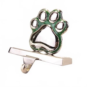 May include: A silver-colored stocking holder with a paw print design. The paw print is outlined in green glitter and has a shiny silver center. The holder has a hook for hanging a stocking.