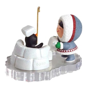 May include: A Christmas ornament featuring a penguin in an igloo and a person in winter clothing. The penguin is black and white, wearing a white square hat. The person is wearing a blue parka with a white fur-lined hood and holding a snowball. The base is clear.