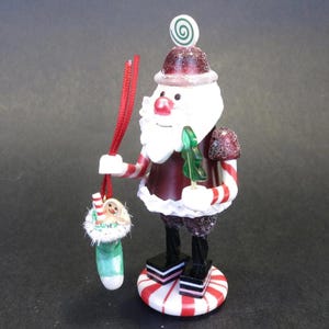 May include: A Santa Claus figurine wearing a dark red hat with a green swirl topper, a matching red coat, and white trim. The figure holds a green tree in one hand and a red string suspending a light green stocking containing a small gingerbread person.