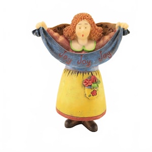 May include: A figurine of a person with curly reddish-brown hair wearing a yellow skirt with a floral pocket and a blue banner draped across their arms. The banner features the repeated red text "Joy Joy Joy" and the figure has a light green collar.