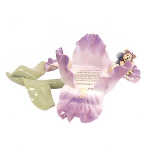 May include: A ceramic candle holder shaped like a purple iris flower with green leaves. A small teddy bear figurine is attached to the edge of the petal. The center of the flower features printed safety text regarding candle usage and fire prevention.