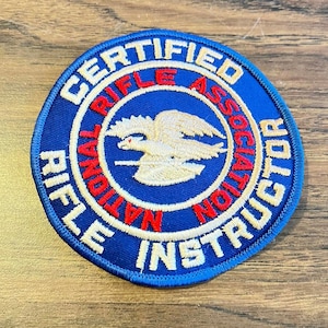 May include: A circular embroidered patch with a blue background and a white border. The text "CERTIFIED RIFLE INSTRUCTOR" is stitched in white, while "NATIONAL RIFLE ASSOCIATION" appears in red. A white eagle emblem is centered in the middle.