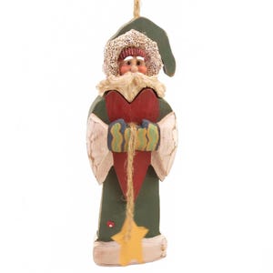 May include: A wooden Christmas ornament in the shape of a figure holding a red heart. The figure has a green coat, a white beard, and a green hat with a white lace trim. A yellow star hangs from the heart.