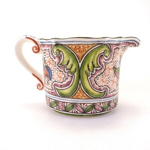May include: A ceramic creamer with a white handle and spout. The creamer is decorated with a floral and leaf pattern in green, pink, and blue. The creamer has a scalloped rim and is approximately 4 inches tall.