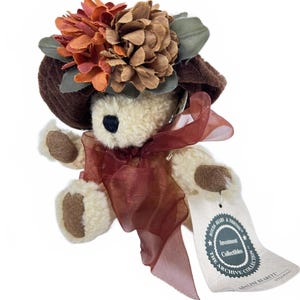 May include: A small, cream-colored teddy bear wearing a brown hat adorned with orange and brown floral accents. The bear has a burgundy ribbon around its neck and a tag that reads "Boyds Bears & Friends".