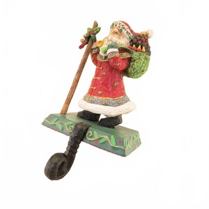 May include: A decorative Christmas stocking holder featuring a figurine of Santa Claus. Santa is dressed in a red coat with gold accents, carrying a sack of gifts and a wooden staff. The base is green with a decorative leaf pattern.