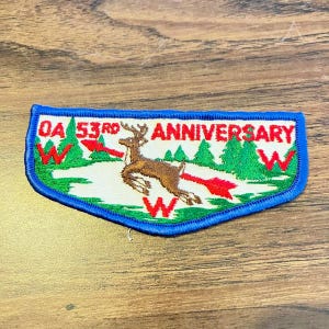 May include: A rectangular embroidered patch with a blue border featuring a brown deer leaping across a white field. The text reads OA 53RD ANNIVERSARY. The design includes green pine trees, red arrows, and red W letters on a white background.