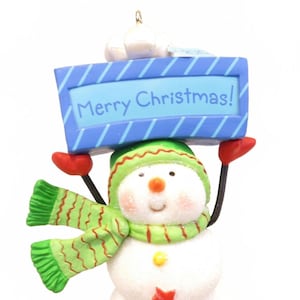 May include: A snowman Christmas ornament with a blue sign that reads "Merry Christmas!". The snowman wears a green hat and scarf, red mittens, and has a white body. The sign is blue with diagonal stripes.