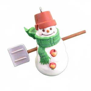May include: A snowman ornament with a red bucket hat, green scarf, and two apple buttons. It holds a shovel and has a smiling face. A festive decoration for the holiday season.