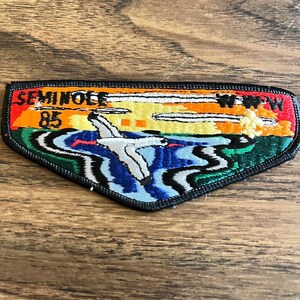 May include: A triangular embroidered patch with a black border featuring a landscape scene with a white bird flying over blue and green water. The sky is orange and yellow with white clouds. Text reads SEMINOLE 85 on the left and W.W.W on the right.