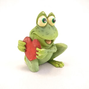 May include: A whimsical green frog figurine with large, expressive eyes and a red heart. The frog is holding the heart with its hands, and the figurine has a textured finish. The frog is a charming decorative item.