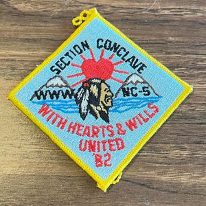 May include: A diamond-shaped patch with a light blue background and yellow border. The patch features a red heart with sun rays, mountains, and text that reads "SECTION CONCLAVE NC-5 WITH HEARTS & WILLS UNITED '82". An illustration of a Native American is also present.