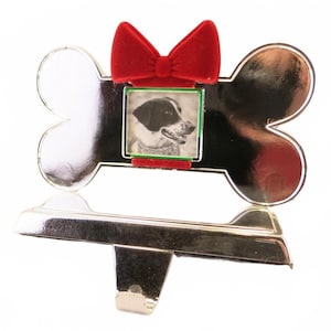 May include: A silver-colored dog bone-shaped stocking holder with a red velvet bow and a small photo frame. The frame holds a black and white photo of a dog. The holder has a hook for hanging.