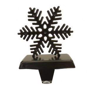 May include: A black metal snowflake stocking holder. The snowflake design is detailed with multiple points and a central circular pattern. The holder has a rectangular base and a hook for hanging stockings. Perfect for holiday decor.