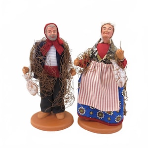 May include: Two ceramic figurines of a fisherman and fisherwoman. The fisherman wears a red scarf, navy jacket, and black pants, with a fishing net draped around him. The fisherwoman wears a red top, patterned shawl, and striped apron. Both hold fish.