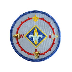May include: Round embroidered patch with a compass design. The patch features a light blue background with a red compass rose, gold directional markers, and a central diamond with a fleur-de-lis. The border is dark blue.