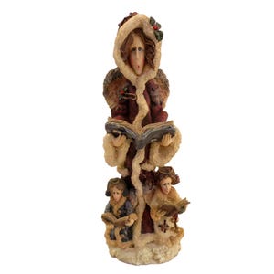 May include: A tall, decorative figurine of carolers. The figurine features a woman in a hooded coat, holding a book, with two smaller figures below. The coats are red and trimmed with faux fur. The figurine is on a white base.
