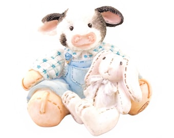 May include: A small figurine of a black and white cow wearing a blue and white checkered shirt and light blue overalls. The cow is sitting down and holding a small white rabbit plush toy with a bow around its neck. The cow has a pink nose.