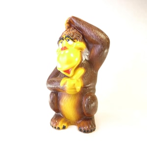 May include: A vintage, brown and yellow gorilla figurine with a surprised expression. The gorilla has a yellow face, hands, and chest, with a red mouth and brown fur. The gorilla is holding its hand to its head.